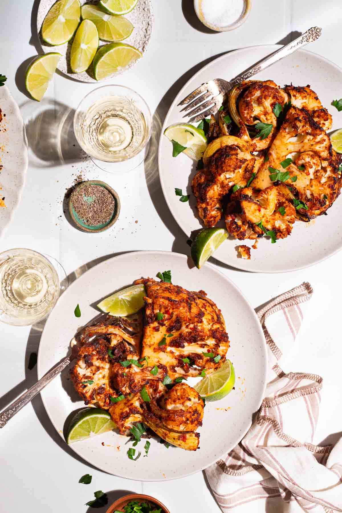 An overhead photo of 2 servings of spicy baked cauliflower topped with chopped parsley and lime wedges on 2 plates with forks. Next to these are 2 glasses of white wine, a plate of lime wedges, bowls of salt and black pepper, and a cloth napkin.