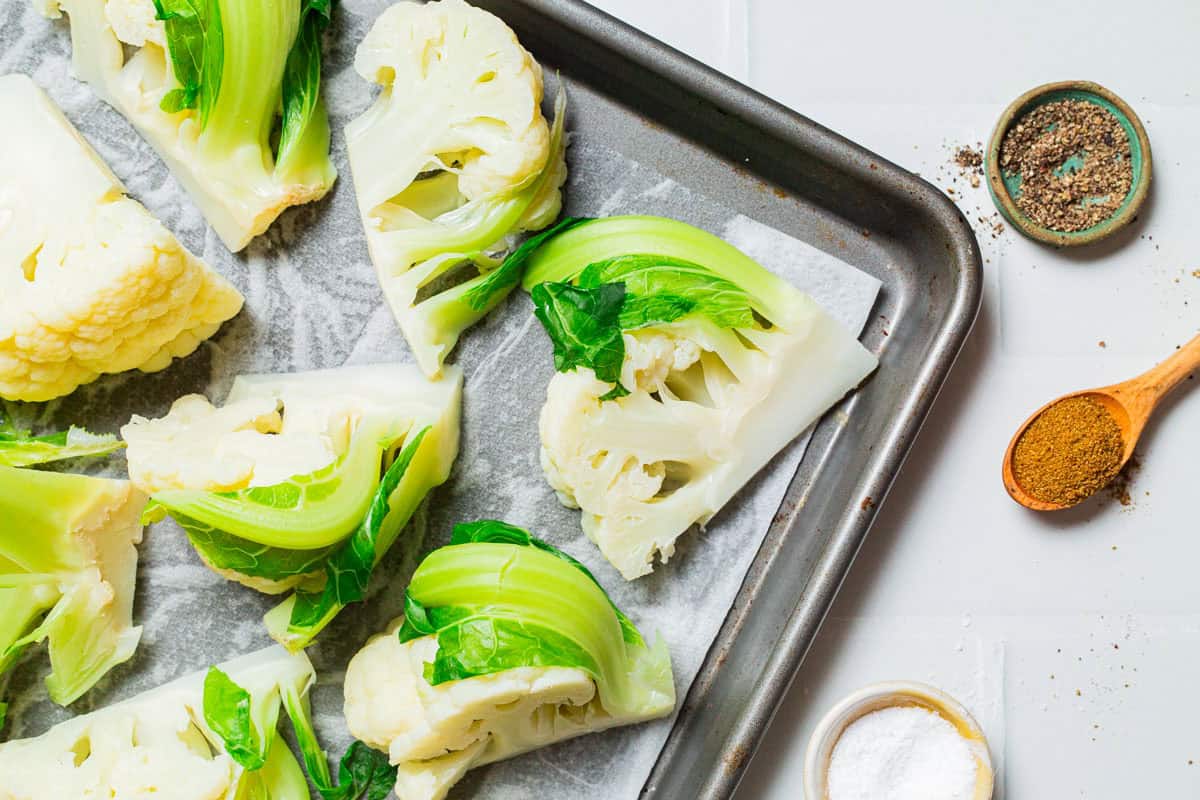 An overhead photo parboiled cauliflower wedges on a paper towel limed sheet pan. Next to this is a wooden spoon with ground cumin, and small bowls of salt and black pepper.