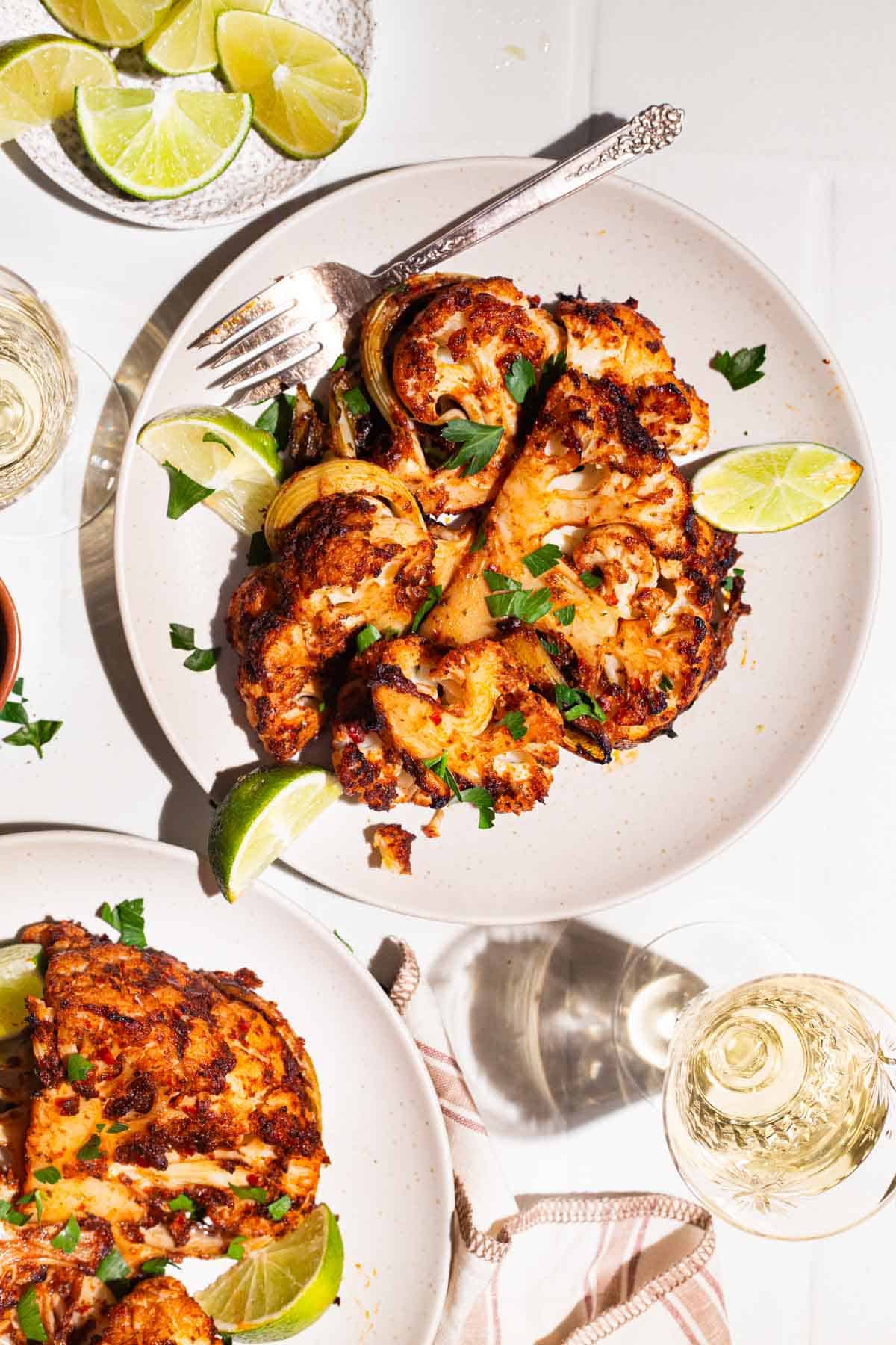 An overhead photo of 2 servings of spicy baked cauliflower topped with chopped parsley and lime wedges on 2 plates, one with a fork. Next to these are 2 glasses of white wine, a plate of lime wedges, and a kitchen towel.