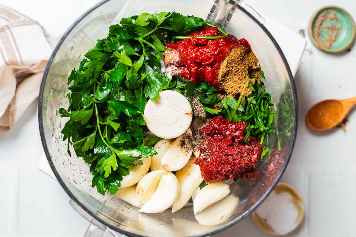 An overhead photo of the ingredients for the garlic harissa sauce in a food processor fitted with a blade just before being mixed together. Next to this is a cloth napkin, bowls of salt and black pepper and a small wooden spoon.