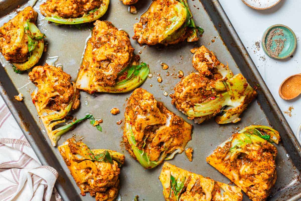 An overhead photo of unbaked spicy cauliflower wedges on a baking sheet. Next to this is a kitchen towel, a bowl of black pepper and a small wooden spoon.