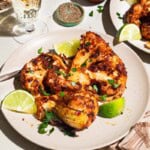 A close up of a serving of spicy baked cauliflower topped with chopped parsley and lime wedges on a plate with a fork. Next to this is a plate with another serving of the cauliflower, a glass of white wine, and a bowl of black pepper and a cloth napkin.