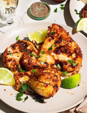 A close up of a serving of spicy baked cauliflower topped with chopped parsley and lime wedges on a plate with a fork. Next to this is a plate with another serving of the cauliflower, a glass of white wine, and a bowl of black pepper and a cloth napkin.