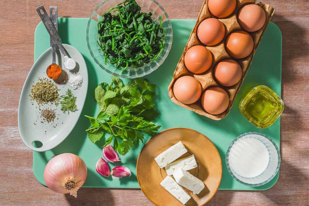 Ingredients for feta spinach frittata including eggs, spinach, oregano, dill, black pepper, paprika, baking powder, salt, onion, parsley, mint, garlic, feta, milk and olive oil.