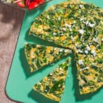 An overhead photo of a feta spinach frittata on a cutting board with two slices cut from it. Next to this is a mediterranean cucumber salad in a bowl.