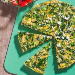 An overhead photo of a feta spinach frittata on a cutting board with two slices cut from it. Next to this is a mediterranean cucumber salad in a bowl.