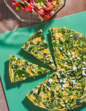 An overhead photo of a feta spinach frittata on a cutting board with two slices cut from it. Next to this is a mediterranean cucumber salad in a bowl with a spoon.