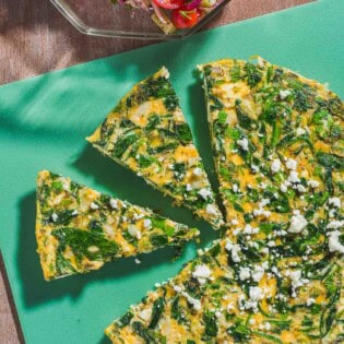 An overhead photo of a feta spinach frittata on a cutting board with two slices cut from it. Next to this is a mediterranean cucumber salad in a bowl with a spoon.