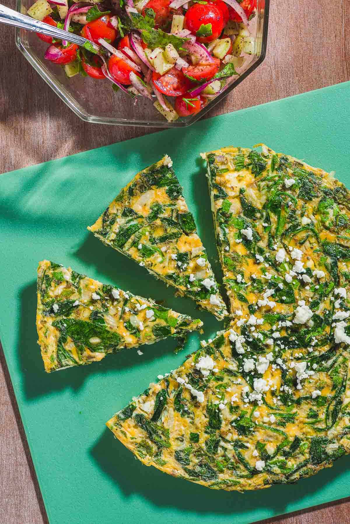 An overhead photo of a feta spinach frittata on a cutting board with two slices cut from it. Next to this is a mediterranean cucumber salad in a bowl with a spoon.