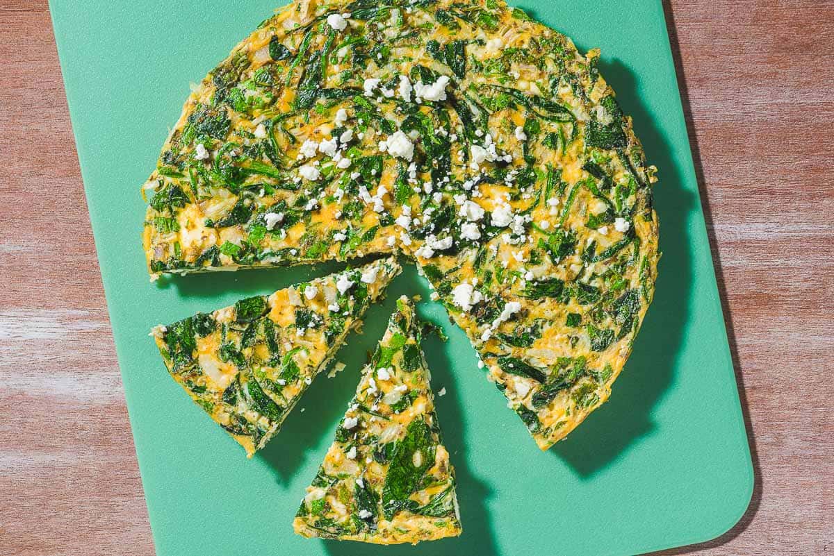 An overhead photo of a feta spinach frittata on a cutting board with two slices cut from it, and one missing.