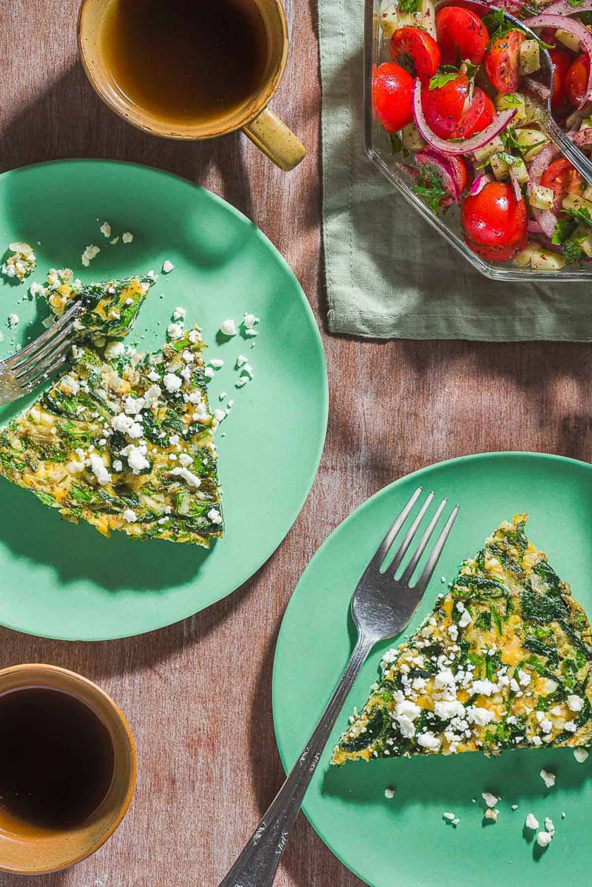 An overhead photo of 2 slices of the feta spinach frittata topped with crumbled feta on two plates with forks. Next to these are 2 cups of coffee and a mediterranean cucumber salad in a bowl with a spoon.