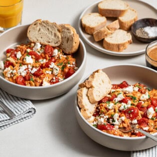 Two bowls of strapastada greek tomato scrambled eggs topped with crumbled feta and chopped parsley, one with a fork. Next to these are 2 cups of espresso, 2 cups of orange juice, a plate of sliced bread with a small bowl of salt and pepper, 2 cloth napkins and a fork.