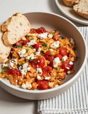 A close up of a serving of strapastada greek tomato scrambled eggs topped with crumbled feta and chopped parsley in a bowl with 2 slices of bread. Next to this is a cloth napkin and a plate of sliced bread.