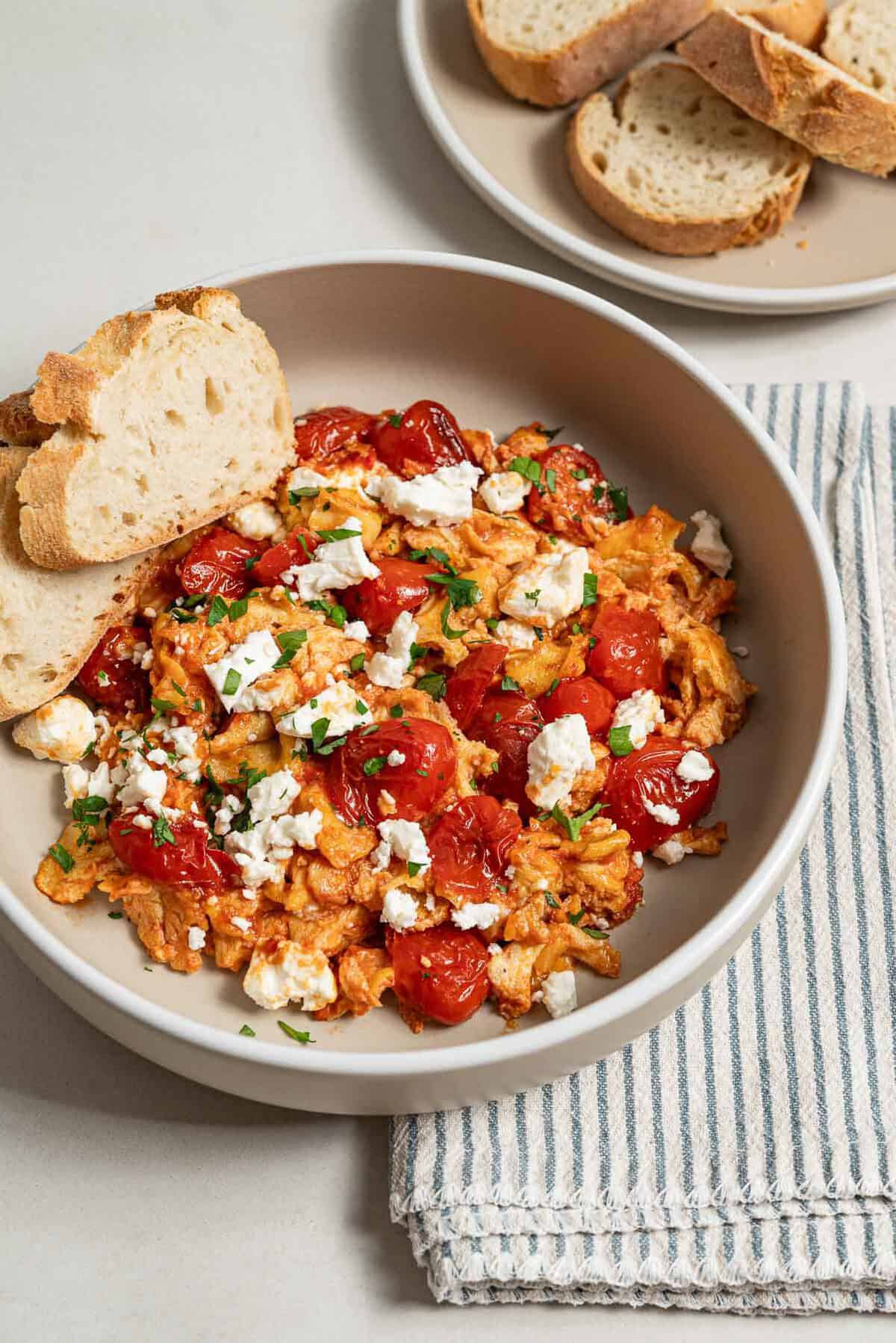 A close up of a serving of strapastada greek tomato scrambled eggs topped with crumbled feta and chopped parsley in a bowl with 2 slices of bread. Next to this is a cloth napkin and a plate of sliced bread.