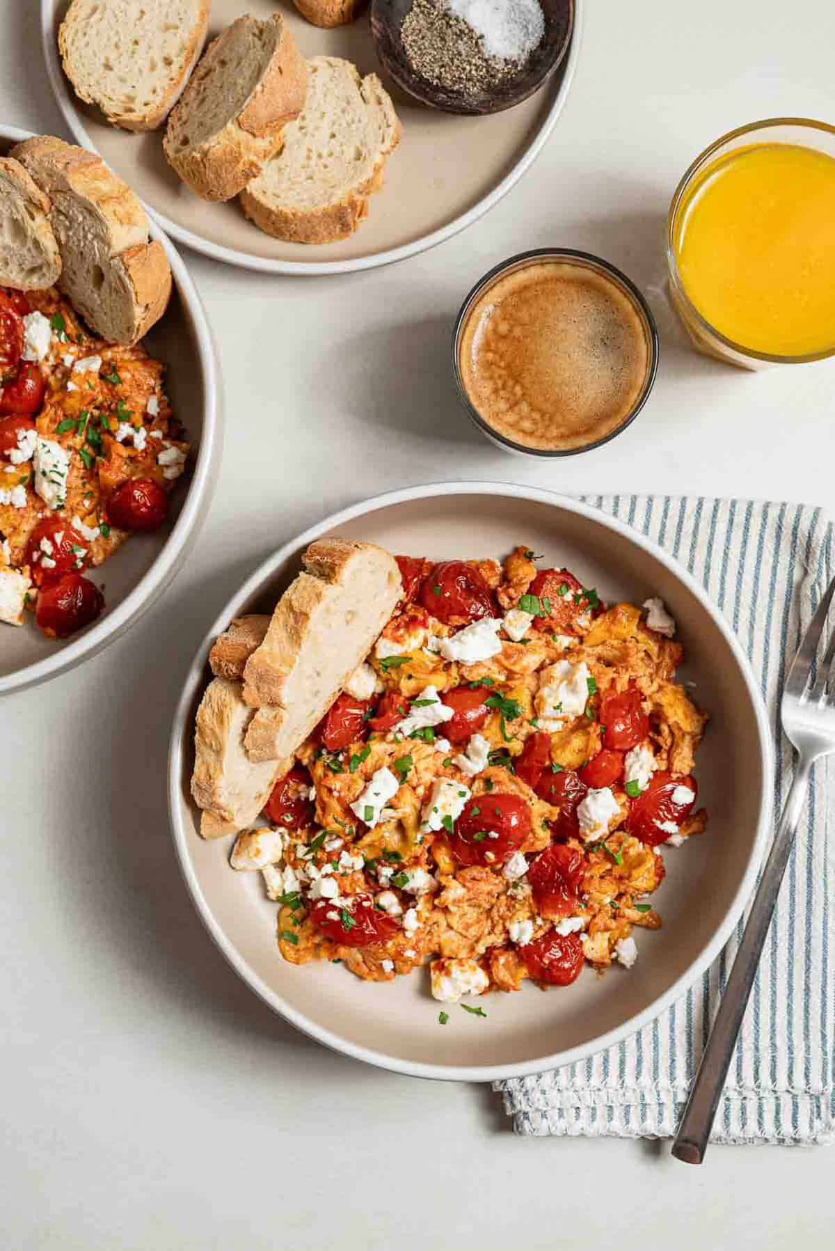 An overhead photo of a serving of strapastada greek tomato scrambled eggs topped with crumbled feta and chopped parsley in a bowl with 2 slices of bread. Next to this is a cloth napkin with a fork, a cup of espresso, a cup of orange juice, a plate of sliced bread, and another bowl of the scrambled eggs.