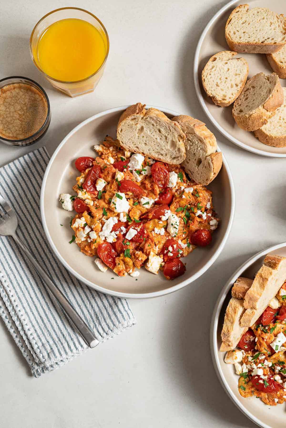 An overhead photo of a serving of strapastada greek tomato scrambled eggs topped with crumbled feta and chopped parsley in a bowl with 2 slices of bread. Next to this is a cloth napkin with a fork, a cup of espresso, a cup of orange juice, a plate of sliced bread, and another bowl of the scrambled eggs.