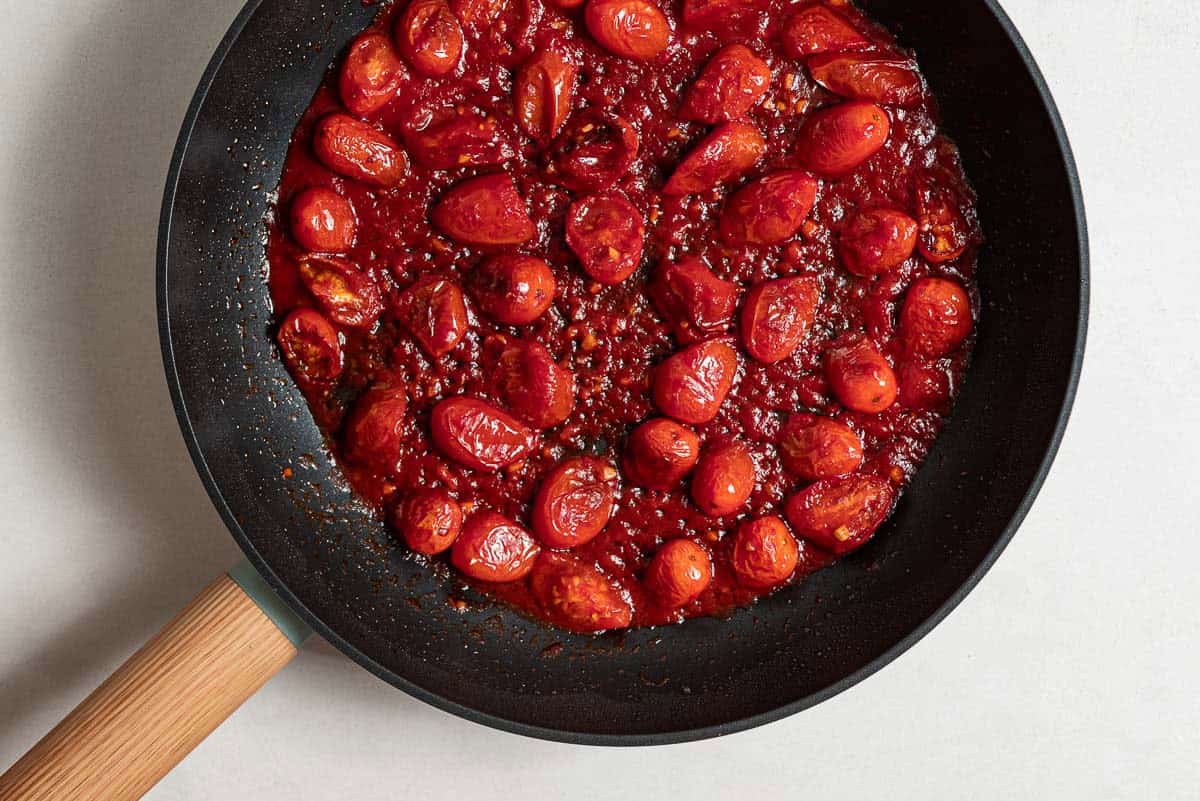 An overhead photo of halved cherry tomatoes simmering with tomato paste, garlic and water in a skillet.