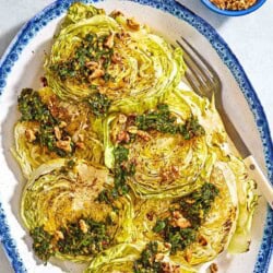 An overhead photo of cabbage steaks topped with green chermoula and toasted walnuts on a serving platter with a fork. Next to this are bowls of chermoula and toasted walnuts.