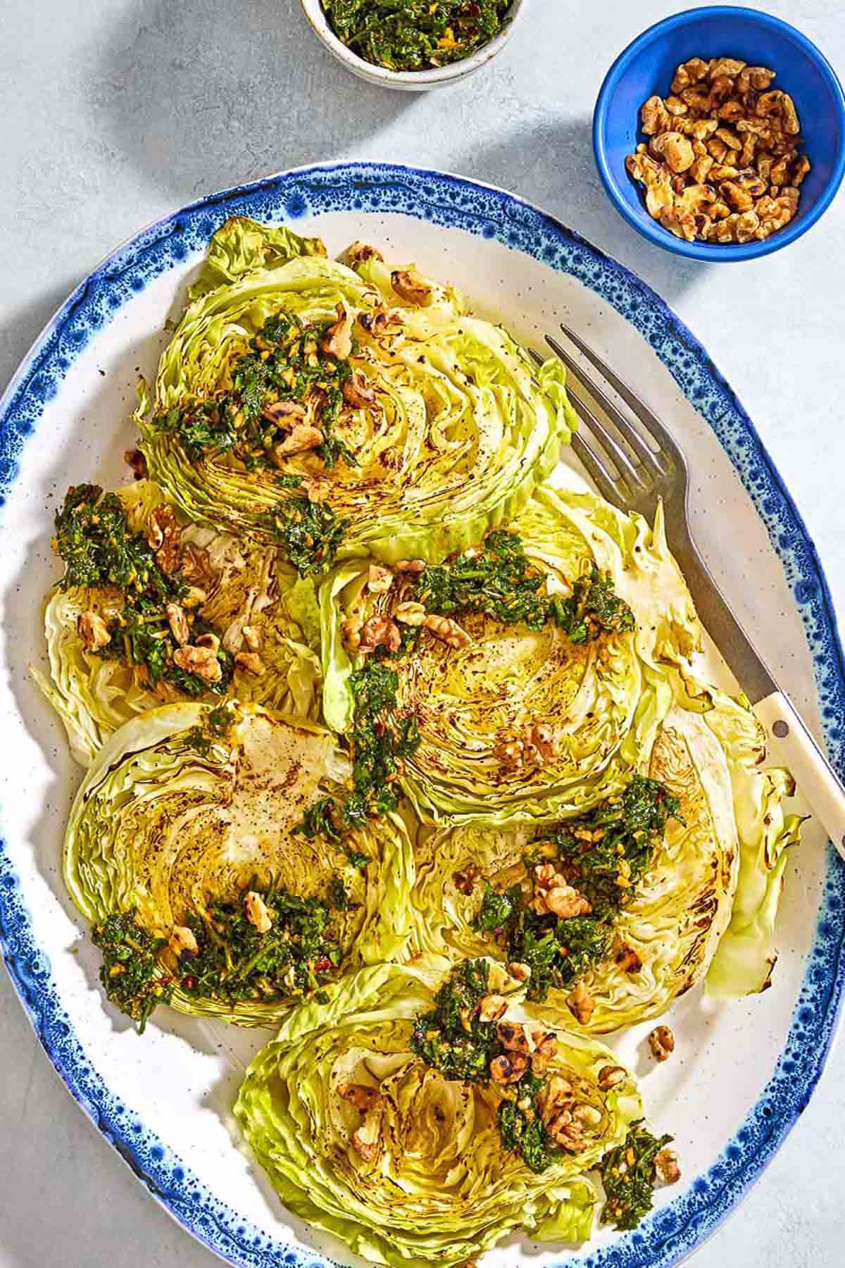 An overhead photo of cabbage steaks topped with green chermoula and toasted walnuts on a serving platter with a fork. Next to this are bowls of chermoula and toasted walnuts.