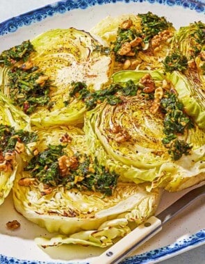 A close up of cabbage steaks topped with green chermoula and toasted walnuts on a serving platter with a fork. Next to this is a bowl of chermoula.