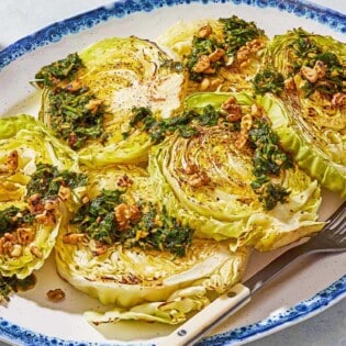 A close up of cabbage steaks topped with green chermoula and toasted walnuts on a serving platter with a fork. Next to this is a bowl of chermoula.