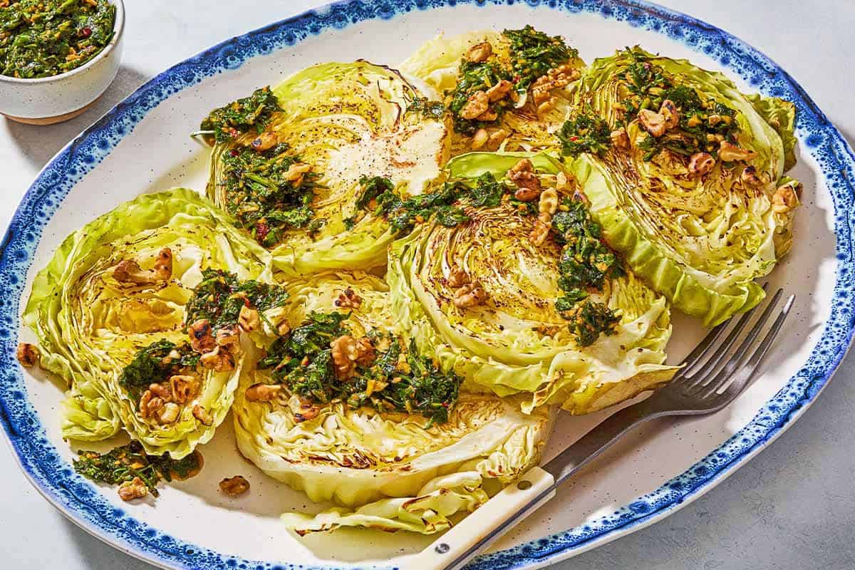 A close up of cabbage steaks topped with green chermoula and toasted walnuts on a serving platter with a fork. Next to this is a bowl of chermoula.