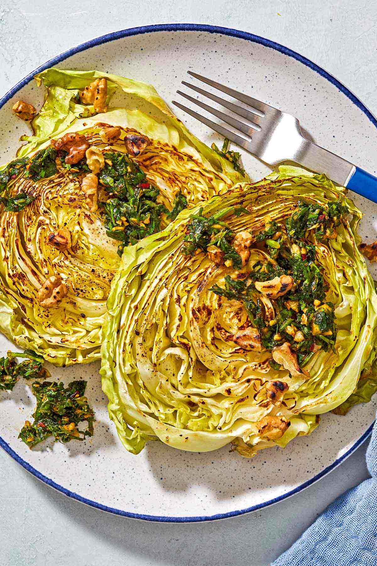 A close up of two cabbage steaks topped with green chermoula and toasted walnuts on a plate with a fork.