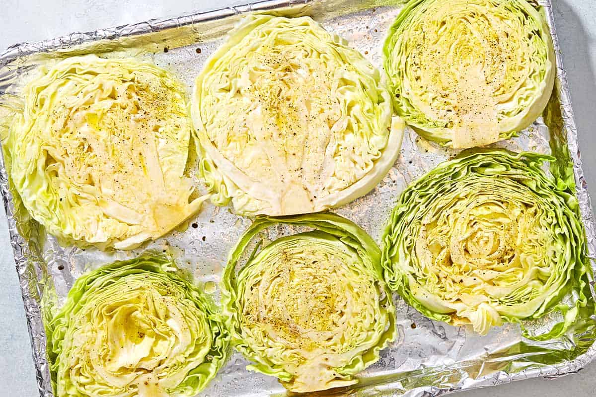 An overhead photo of uncooked cabbage steaks seasoned with salt and pepper on a foil-lined baking sheet.