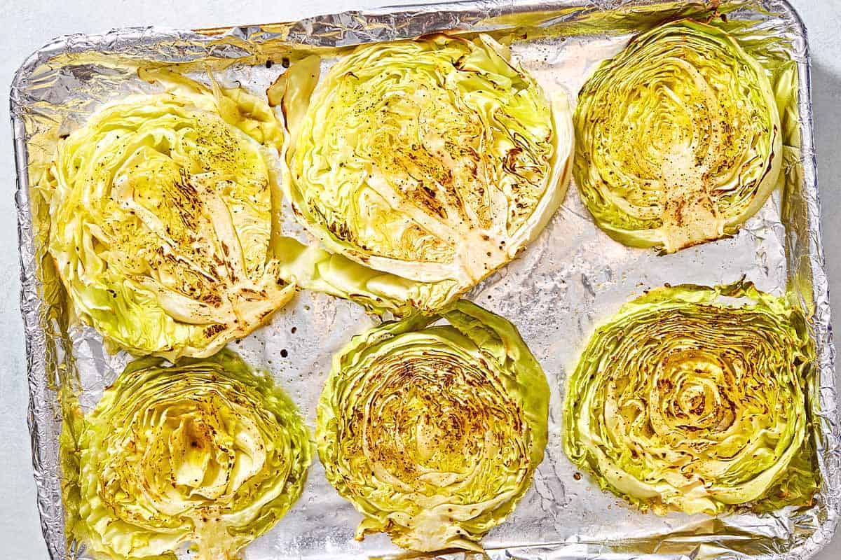 An overhead photo of cooked cabbage steaks seasoned with salt and pepper on a foil-lined baking sheet.