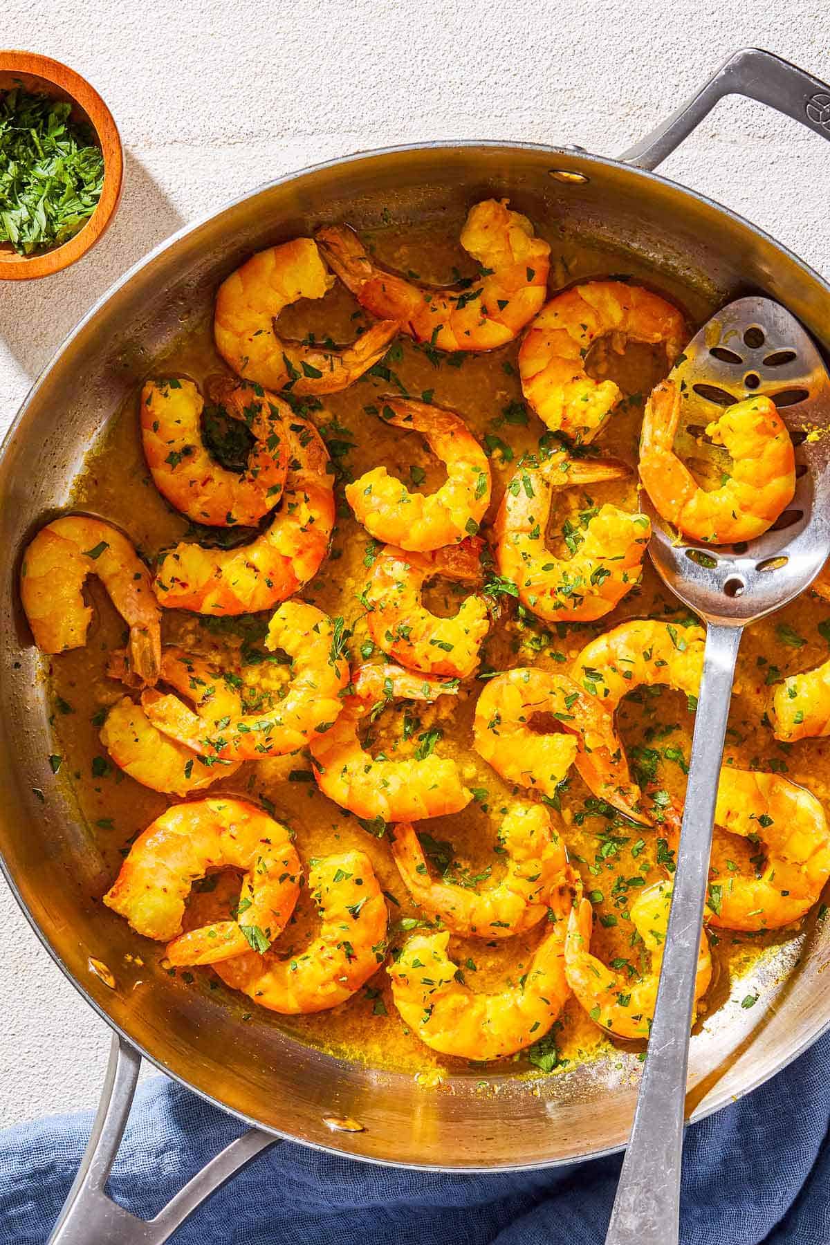 An overhead photo of poached shrimp in a saffron and white wine sauce in a skillet with a spoon. Next to this is a bowl of chopped parsley and a cloth napkin.