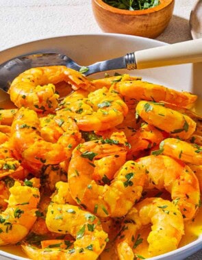 A close up of poached shrimp in a saffron and white wine sauce in a bowl with a spoon. Next to this is a bowl of chopped parsley, a cloth napkin, and some crusty bread on a plate.