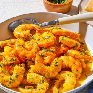 A close up of poached shrimp in a saffron and white wine sauce in a bowl with a spoon. Next to this is a bowl of chopped parsley, a cloth napkin, and some crusty bread on a plate.