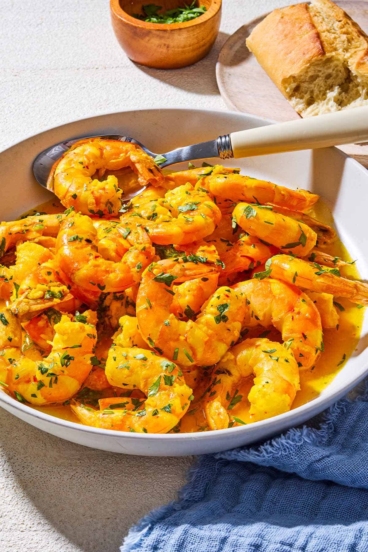 A close up of poached shrimp in a saffron and white wine sauce in a bowl with a spoon. Next to this is a bowl of chopped parsley, a cloth napkin, and some crusty bread on a plate.