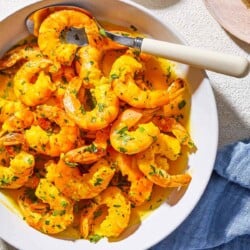 An overhead photo of poached shrimp in a saffron and white wine sauce in a bowl with a spoon. Next to this is a bowl of chopped parsley and a cloth napkin.