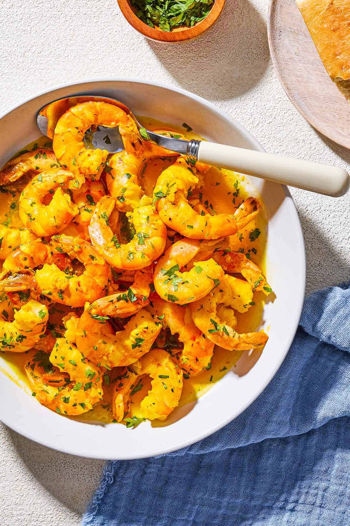 An overhead photo of poached shrimp in a saffron and white wine sauce in a bowl with a spoon. Next to this is a bowl of chopped parsley and a cloth napkin.