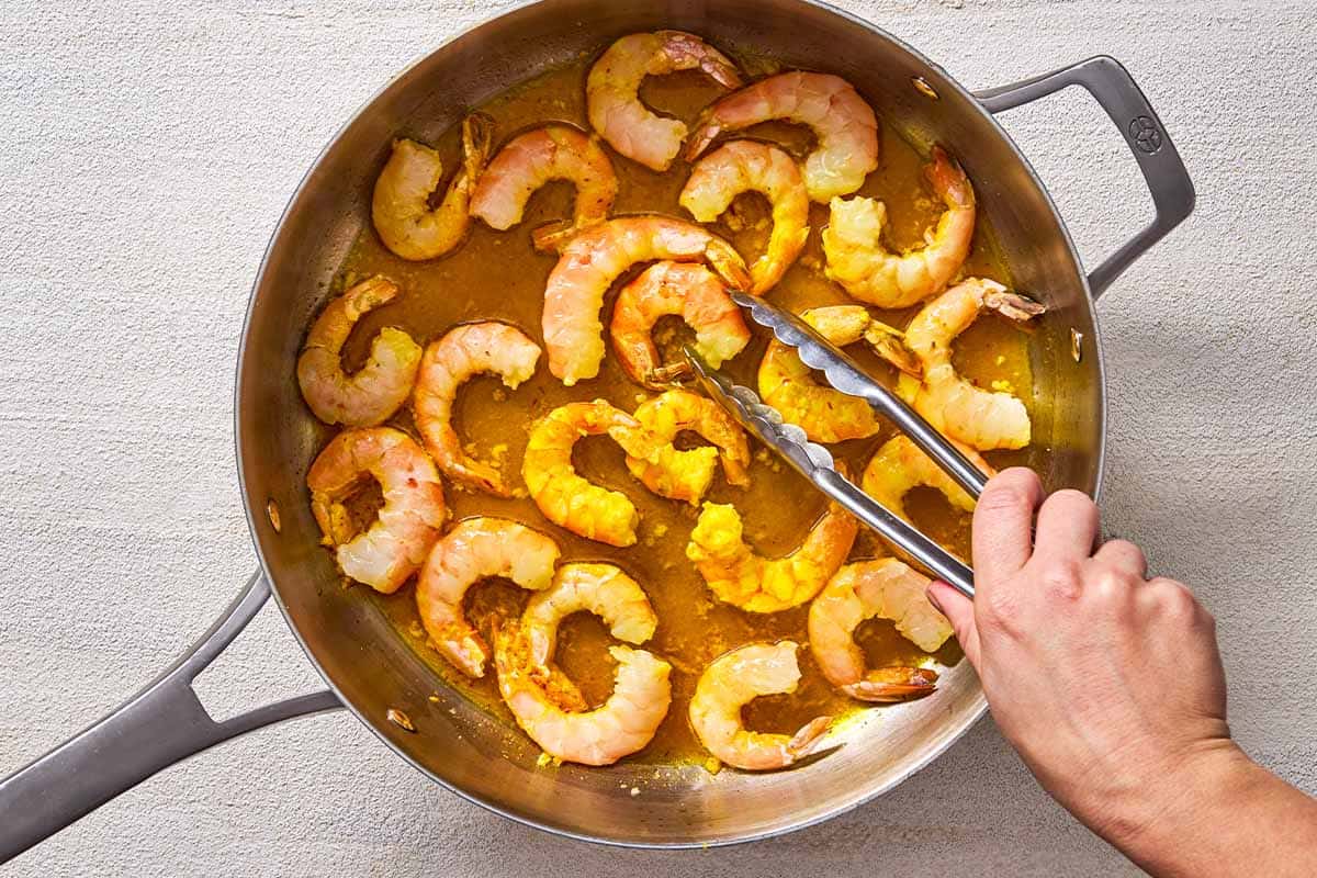 An overhead photo of shrimp being poached in a skillet with a pair of tongs.