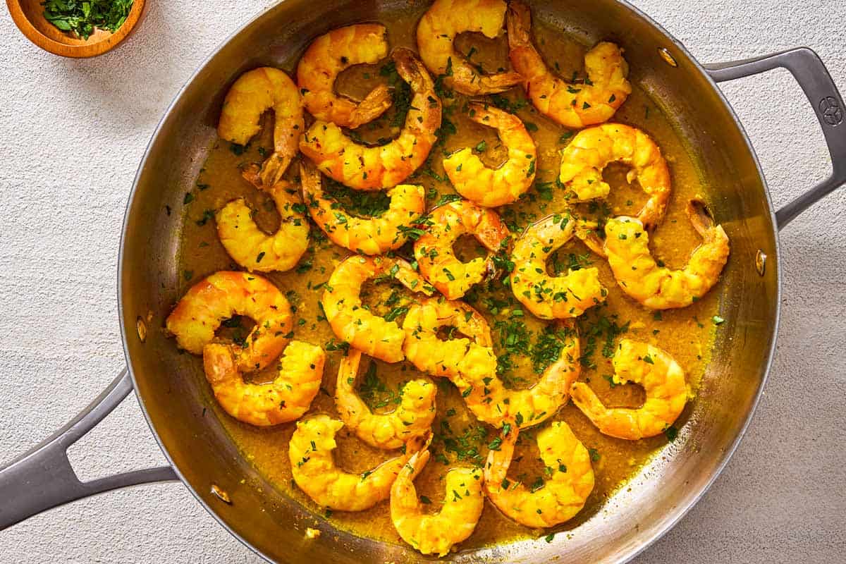 An overhead photo of poached shrimp in a saffron and white wine sauce in a skillet next to a bowl of chopped parsley.