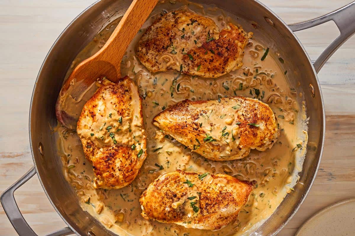 An overhead photo of 4 chicken breast halves and the tarragon mustard sauce in a skillet with a wooden spoon.