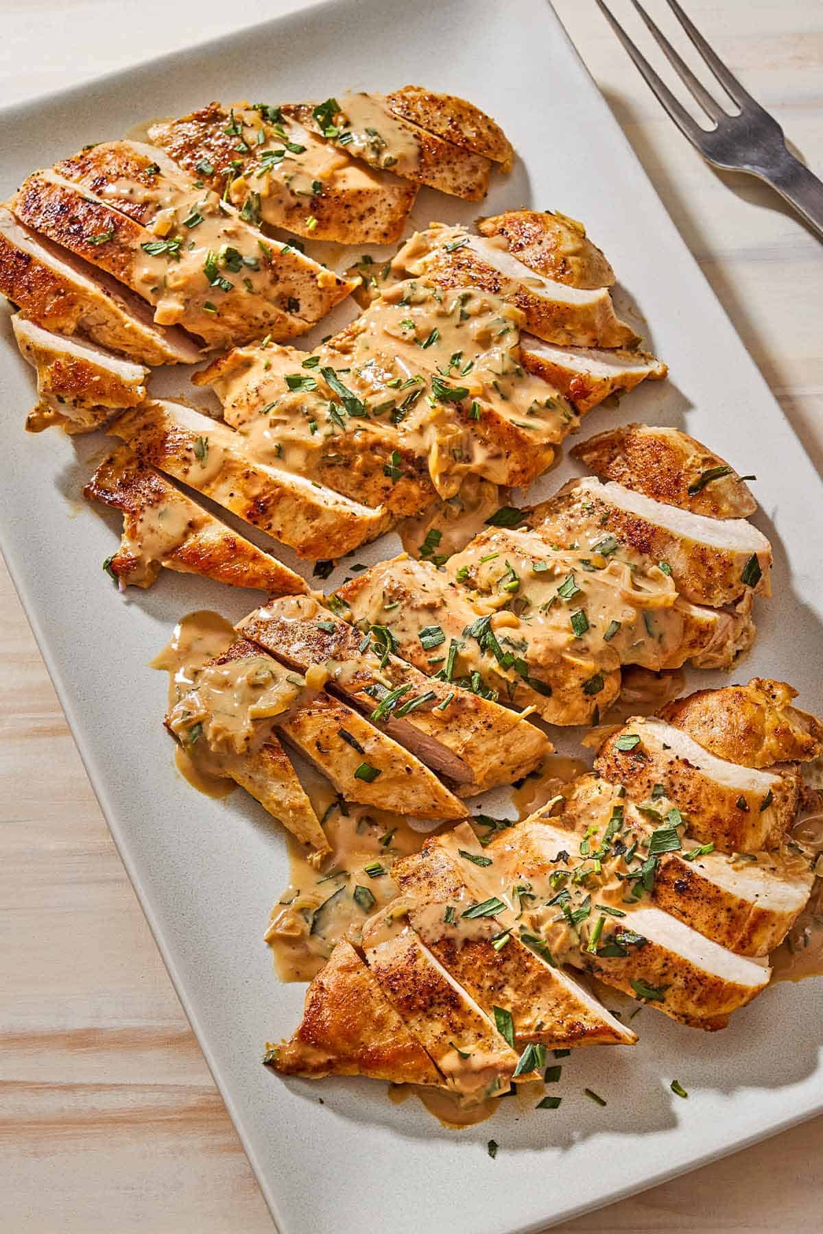 An overhead photo of 4 sliced chicken breasts topped with tarragon mustard sauce on a platter next to a serving fork.