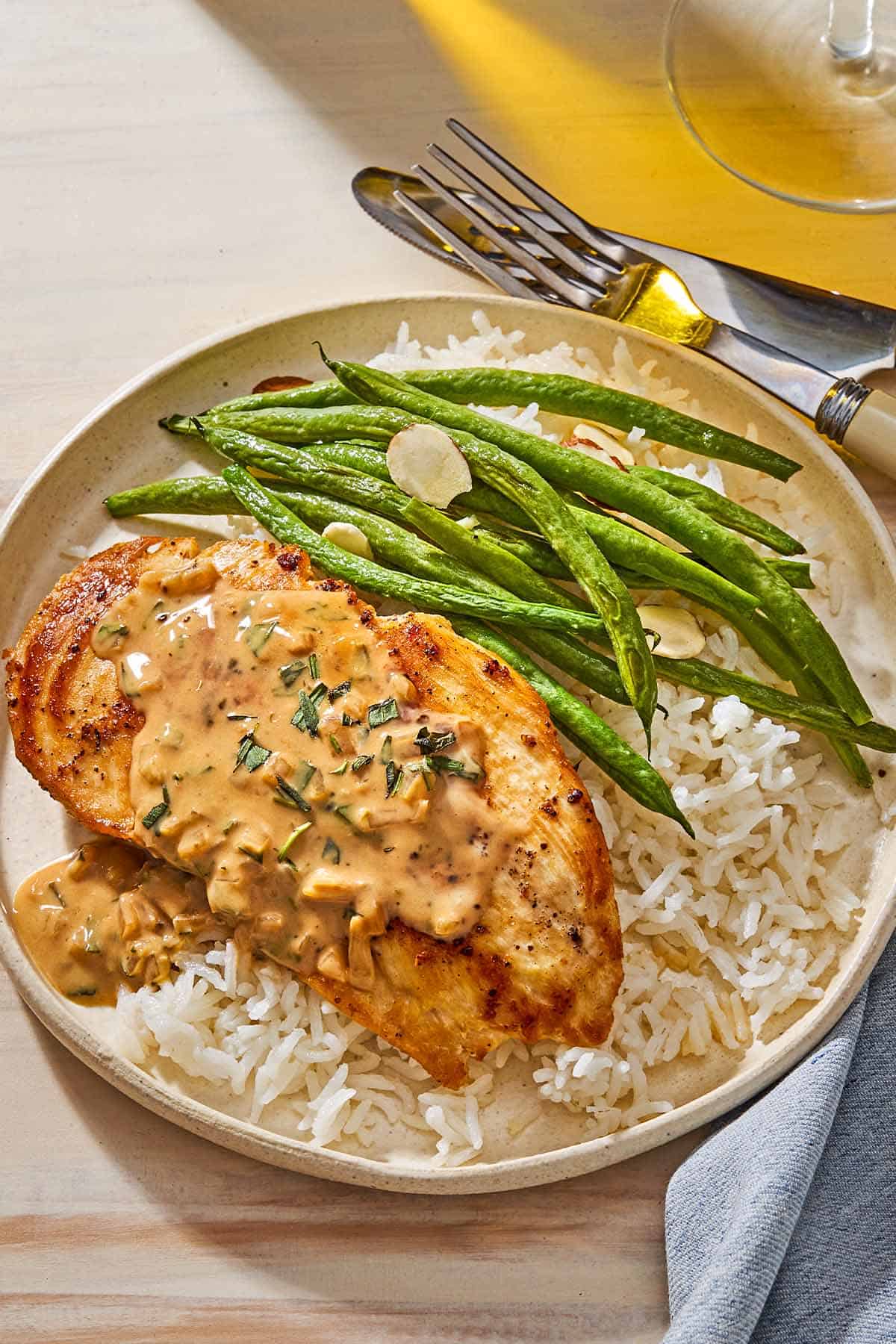 A serving of chicken with tarragon mustard sauce and green beans on a bed of rice on a plate. Next to this is a cloth napkin, a knife and a fork.