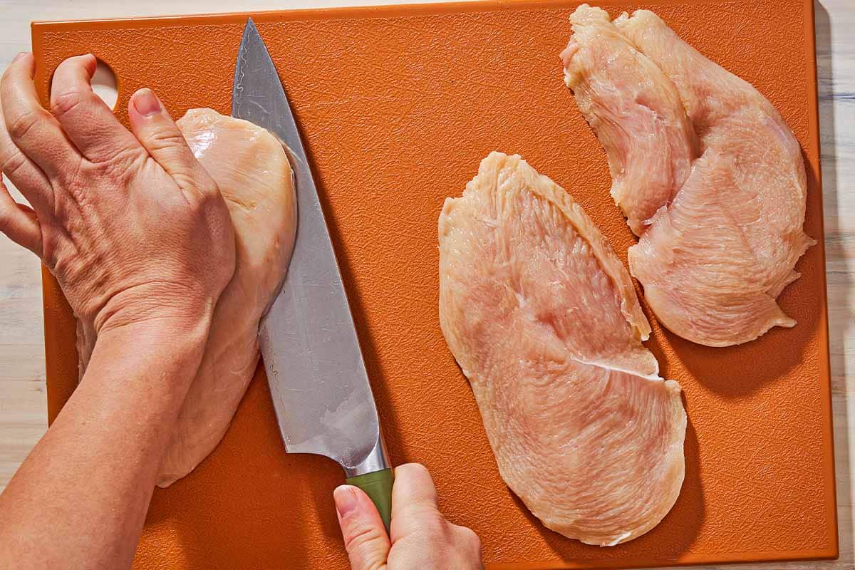 An overhead photo of a whole chicken breast being cut in half with a knife. Next to this on the cutting board is 2 chicken breast halves.