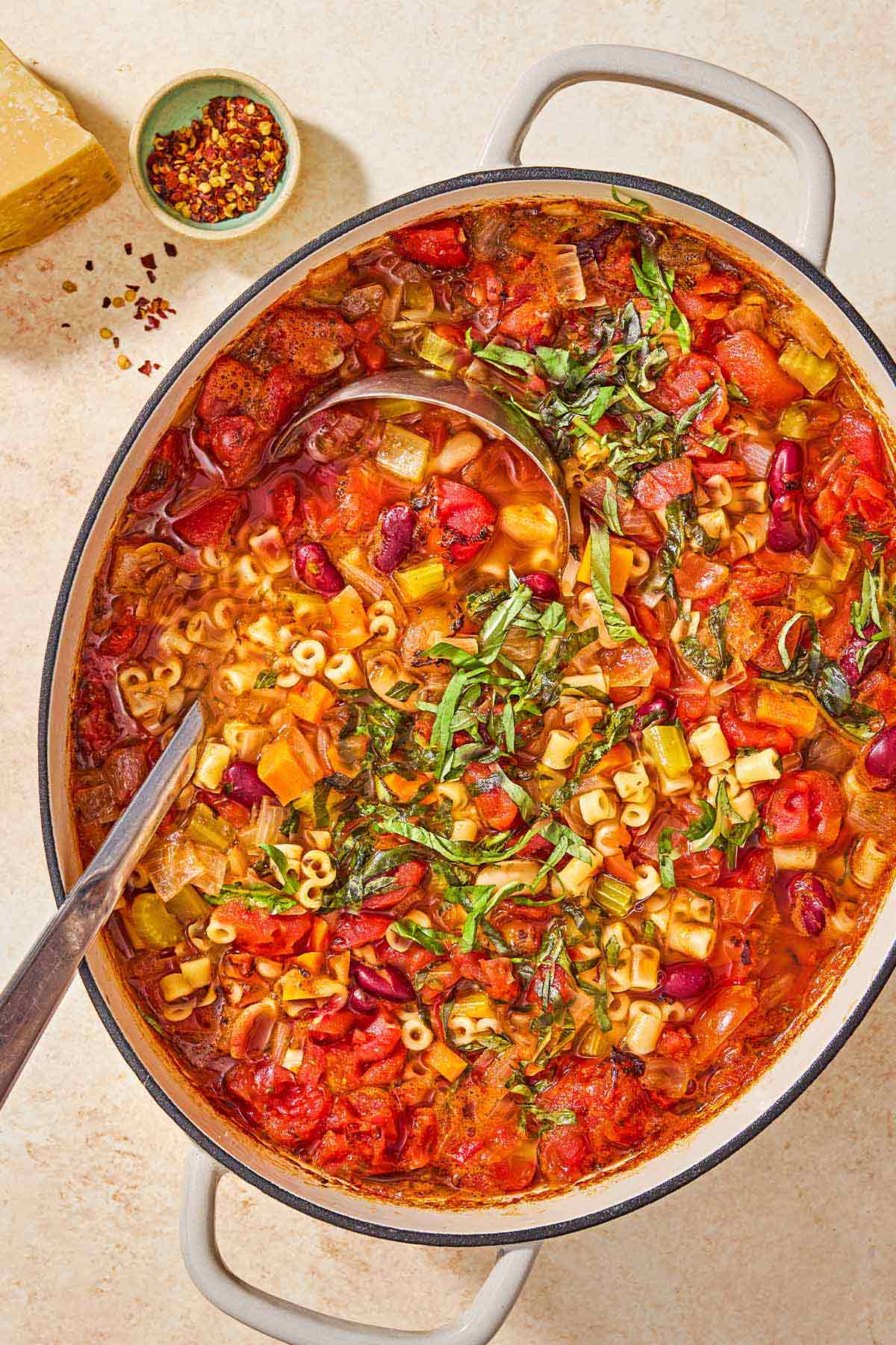 An overhead photo of a pot of pasta fagioli garnished with fresh basil with a ladle. Next to this is a small bowl of red pepper flakes.