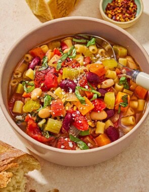 A close up of a bowl of pasta fagioli with a spoon. Next to this is a small bowl of red pepper flakes, a wedge of parmesan cheese, and a piece of crusty bread.