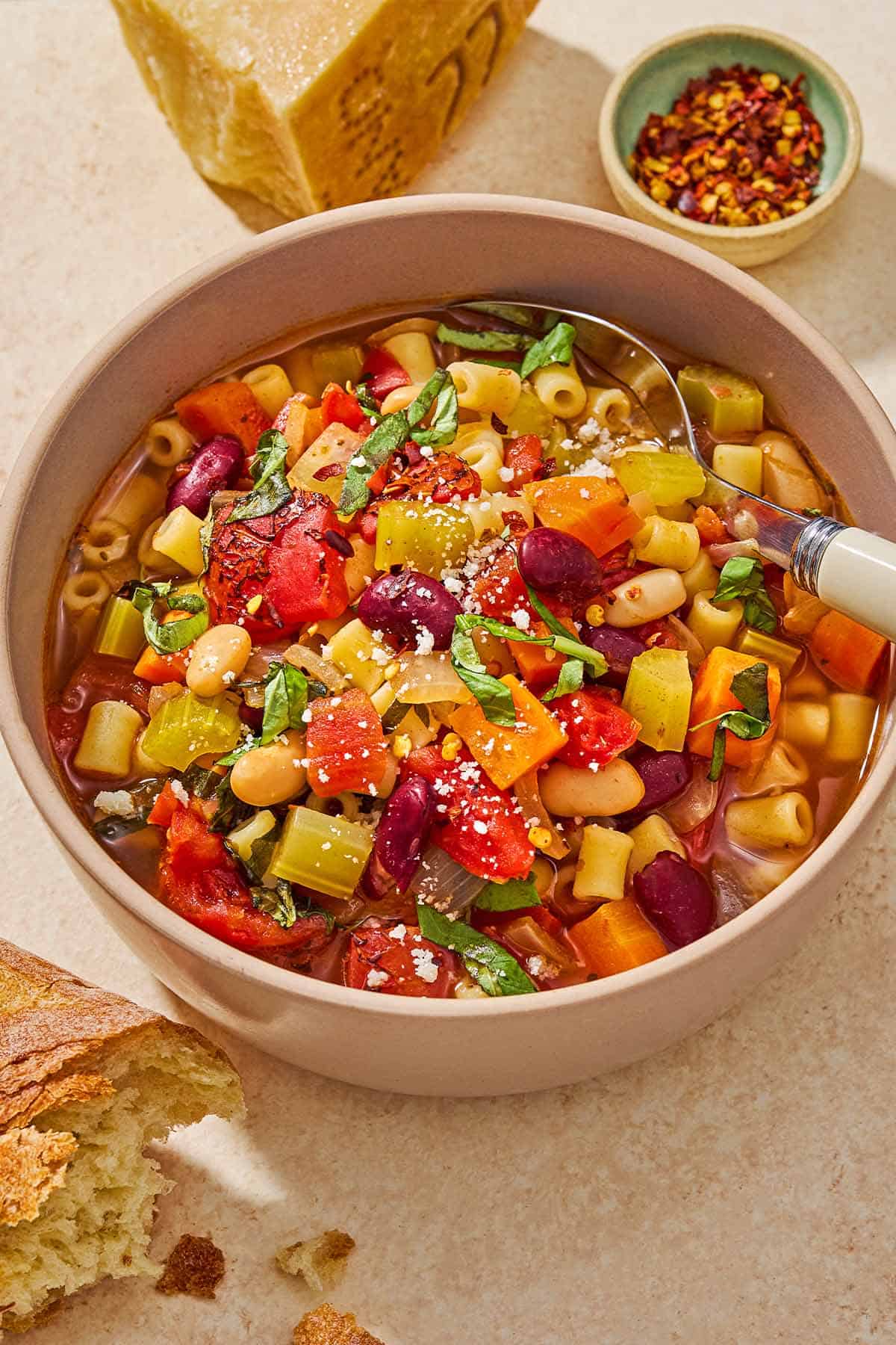 A close up of a bowl of pasta fagioli with a spoon. Next to this is a small bowl of red pepper flakes, a wedge of parmesan cheese, and a piece of crusty bread.