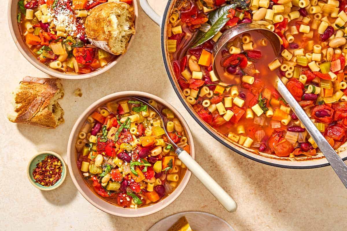 An overhead photo of a bowl of pasta fagioli with a spoon. Next to this is the rest of the pasta in a pot, another bowl of the pasta with a piece of crusty bread in it, a small bowl of red pepper flakes, and another piece of crusty bread.