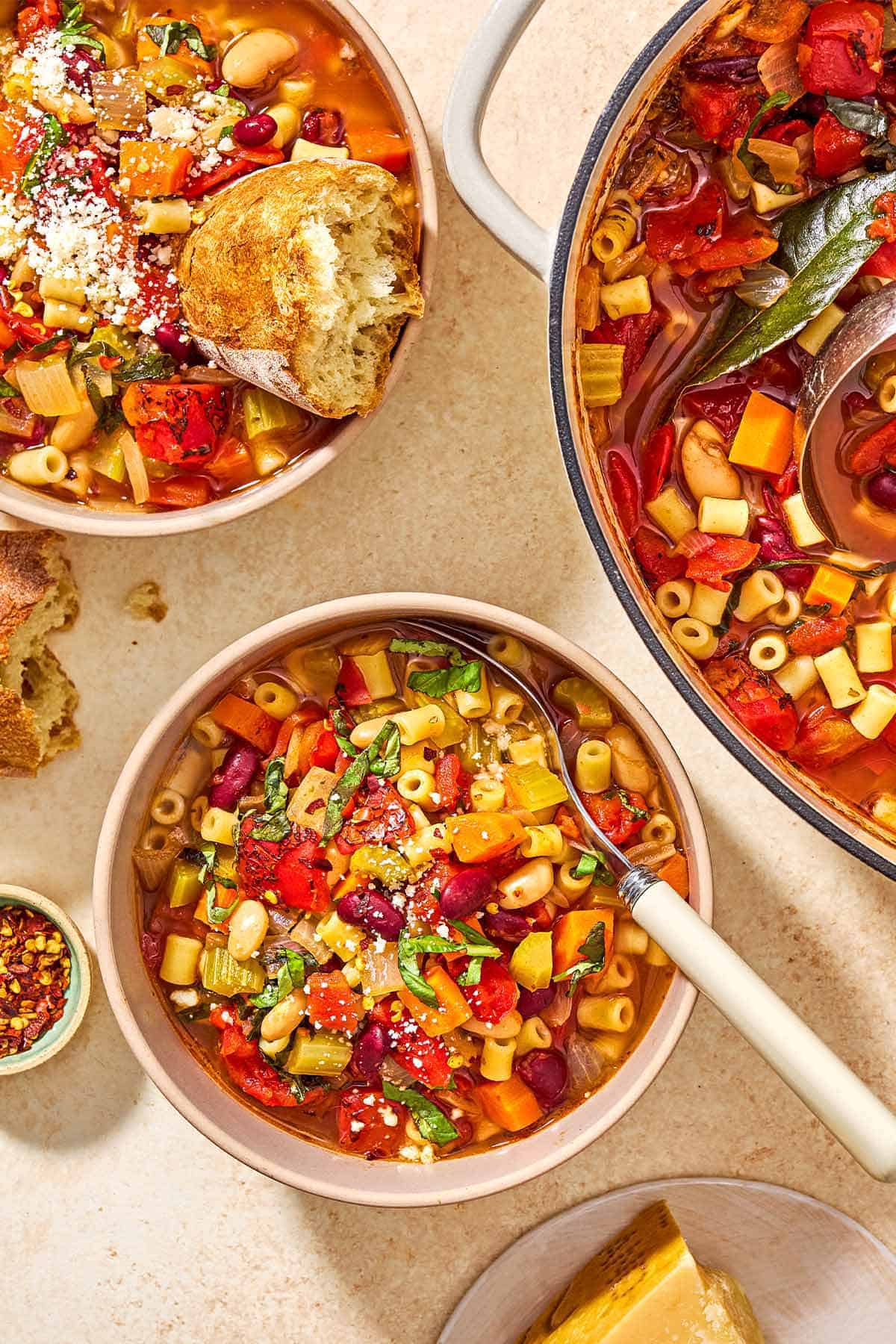 An overhead photo of a bowl of pasta fagioli with a spoon. Next to this is the rest of the pasta in a pot, another bowl of the pasta with a piece of crusty bread in it, a small bowl of red pepper flakes, and another piece of crusty bread.