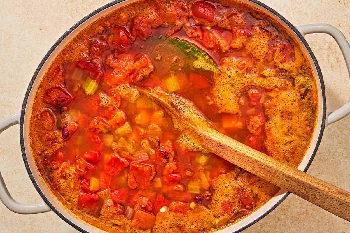 An overhead photo of the vegetables, diced tomatoes, broth, and bean simmering together in a pot with a wooden spoon.