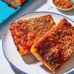 A close up of 2 slices of sfincione sicilian pizza on a plate. Next to this is the rest of the pizza on a sheet pan and a small bowl of red pepper flakes.