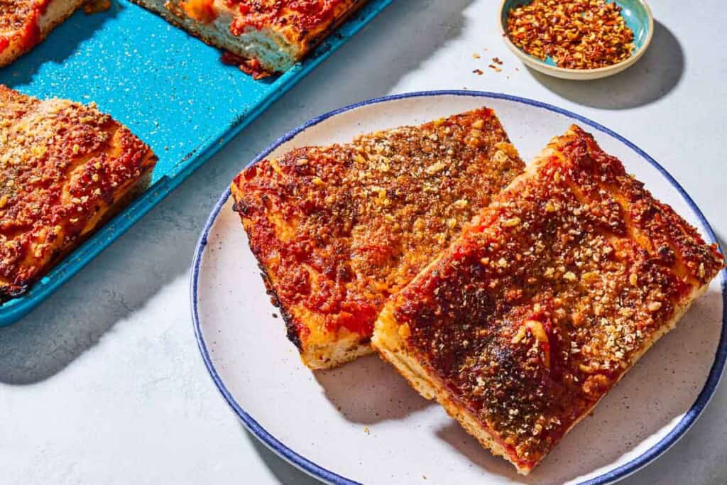 A close up of 2 slices of sfincione sicilian pizza on a plate. Next to this is the rest of the pizza on a sheet pan and a small bowl of red pepper flakes.