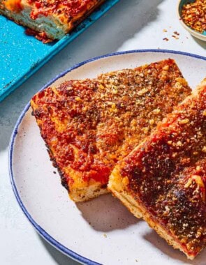 A close up of 2 slices of sfincione sicilian pizza on a plate. Next to this is the rest of the pizza on a sheet pan and a small bowl of red pepper flakes.